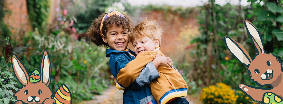 A wide-shot photograph shows two young children, a boy and a girl, hugging and smiling on a winding path through a garden. The girl wears a blue jacket and the boy wears a mustard yellow jacket. The image has cartoon illustrations of Easter bunnies on either side.